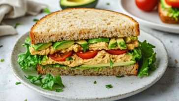 Vegan Sandwich With Avocado, Tomatoes, Lettuce, And Hummus Cut In Half On A White Plate