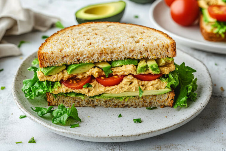 Vegan Sandwich With Avocado, Tomatoes, Lettuce, And Hummus Cut In Half On A White Plate